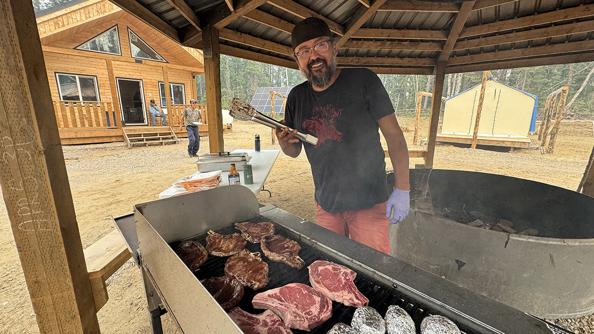 Un homme barbu sourit pendant qu’il cuisine sur le gril dans un pavillon. Une tente et un grand chalet sont visibles en arrière-plan.