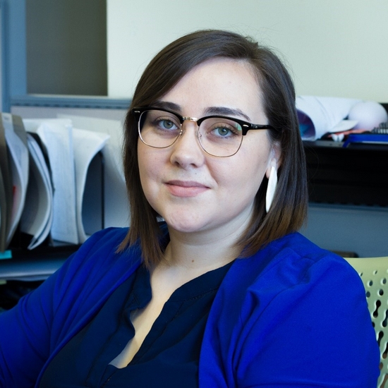 Joy seated at a desk in an office, wearing glasses and a blue blazer, with organized documents and office supplies in the background.